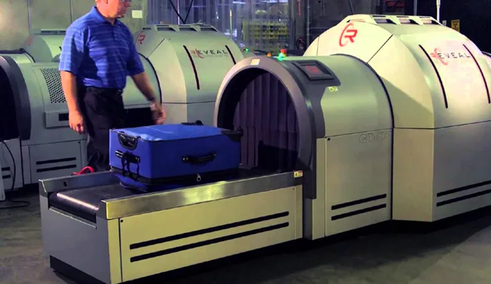 A blue suitcase moves through an X-ray scanner, part of air cargo operations. A worker stands nearby.